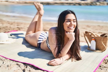 Téléchargez les photos : Jeune femme hispanique portant un bikini allongé sur le sable au bord de la mer - en image libre de droit