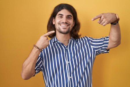 Téléchargez les photos : Homme hispanique avec les cheveux longs debout sur fond jaune souriant gai montrant et pointant avec les doigts dents et la bouche. concept de santé dentaire. - en image libre de droit