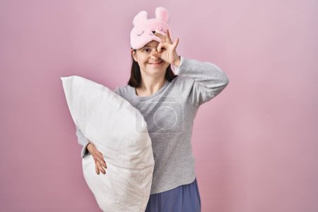 Photo for Woman with down syndrome wearing sleeping mask hugging pillow doing ok gesture with hand smiling, eye looking through fingers with happy face. - Royalty Free Image