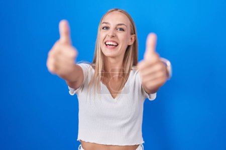Photo for Young caucasian woman standing over blue background approving doing positive gesture with hand, thumbs up smiling and happy for success. winner gesture. - Royalty Free Image
