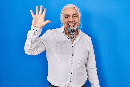 Téléchargez les photos : Homme d'âge moyen avec les cheveux gris debout sur fond bleu montrant et pointant vers le haut avec les doigts numéro cinq tout en souriant confiant et heureux. - en image libre de droit