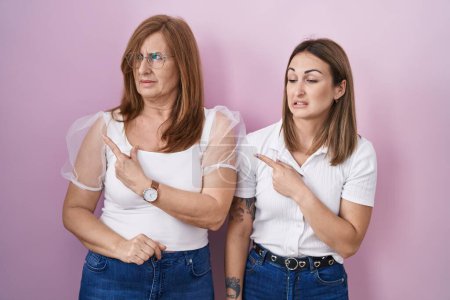 Foto de Madre e hija hispanas vistiendo camiseta blanca casual sobre fondo rosa señalando preocupadas y nerviosas con el dedo índice, expresión preocupada y sorprendida - Imagen libre de derechos