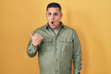 Photo for Hispanic young man standing over yellow background surprised pointing with hand finger to the side, open mouth amazed expression. - Royalty Free Image