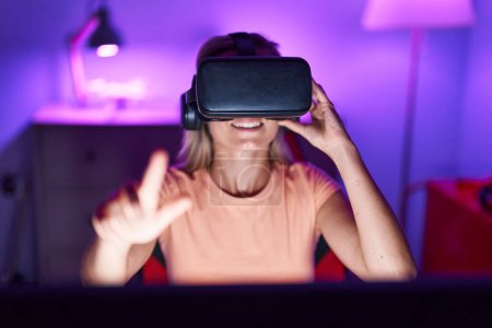 Téléchargez les photos : Jeune femme blonde streamer jouer à un jeu vidéo en utilisant des lunettes de réalité virtuelle à la salle de jeu - en image libre de droit