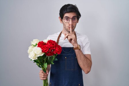 Photo for Young hispanic man holding bouquet of white and red roses asking to be quiet with finger on lips. silence and secret concept. - Royalty Free Image