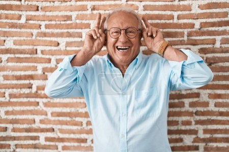 Téléchargez les photos : Homme âgé avec les cheveux gris debout sur le mur de briques posant drôle et fou avec les doigts sur la tête comme des oreilles de lapin, souriant joyeux - en image libre de droit