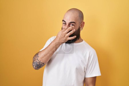 Téléchargez les photos : Jeune homme hispanique avec barbe et tatouages debout sur fond jaune sentant quelque chose de puant et dégoûtant, odeur intolérable, retenant souffle avec les doigts sur le nez. mauvaise odeur - en image libre de droit