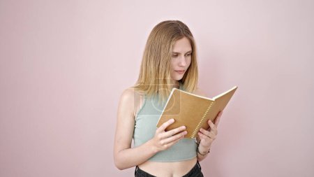Téléchargez les photos : Jeune femme blonde lisant un livre avec un visage sérieux sur fond rose isolé - en image libre de droit