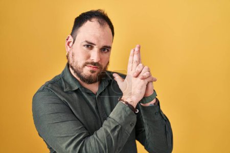 Foto de Hombre hispano de talla grande con barba de pie sobre fondo amarillo sosteniendo un arma simbólica con gesto de mano, jugando a matar armas de fuego, cara enojada - Imagen libre de derechos