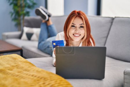 Téléchargez les photos : Jeune femme caucasienne utilisant ordinateur portable et carte de crédit couché sur le canapé à la maison - en image libre de droit