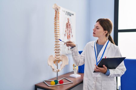 Foto de Joven pisioterapeuta rubia señalando modelo anatómico de columna vertebral sujetando portapapeles en clínica de rehabilitación - Imagen libre de derechos