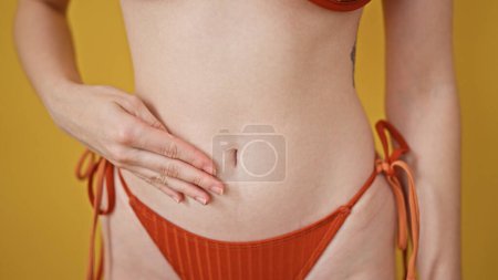 Foto de Mujer rubia joven con bikini que masajea el vientre sobre un fondo amarillo aislado - Imagen libre de derechos