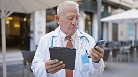 Foto de Senior doctor hombre de pelo gris usando touchpad y teléfono inteligente en la terraza de la cafetería - Imagen libre de derechos