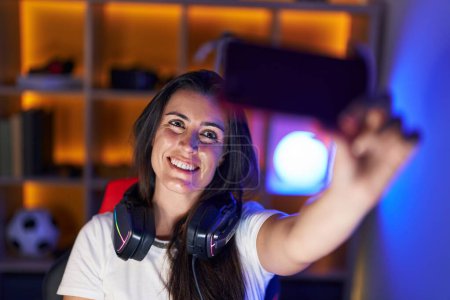Foto de Young beautiful hispanic woman streamer smiling confident make selfie by smartphone at gaming room - Imagen libre de derechos