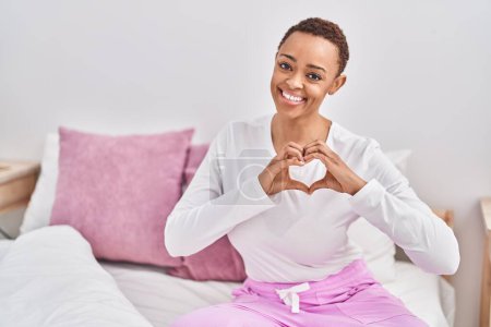 Photo for African american woman doing heart gesture sitting on bed at bedroom - Royalty Free Image