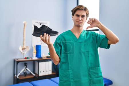 Photo for Young man working at physiotherapy clinic holding shoe with angry face, negative sign showing dislike with thumbs down, rejection concept - Royalty Free Image