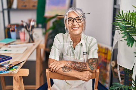 Foto de Mujer artista de pelo gris de mediana edad sonriendo confiada sentada con los brazos cruzados gesto en el estudio de arte - Imagen libre de derechos