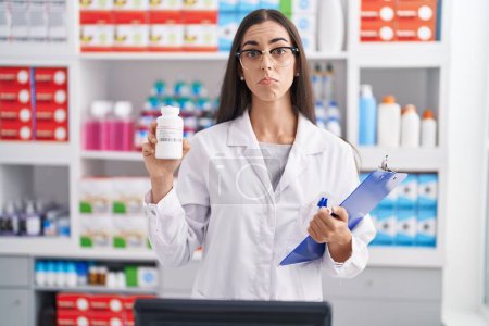 Photo for Young brunette woman working at pharmacy drugstore holding pills depressed and worry for distress, crying angry and afraid. sad expression. - Royalty Free Image