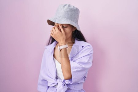 Foto de Mujer hispana joven de pie sobre fondo rosa usando sombrero cansado frotando nariz y ojos sintiendo fatiga y dolor de cabeza. concepto de estrés y frustración. - Imagen libre de derechos