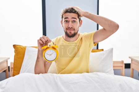 Foto de Hombre hispano con barba sosteniendo el despertador en la cama estresado y frustrado con la mano en la cabeza, rostro sorprendido y enojado - Imagen libre de derechos