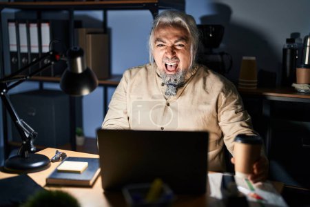 Photo for Middle age man with grey hair working at the office at night angry and mad screaming frustrated and furious, shouting with anger. rage and aggressive concept. - Royalty Free Image