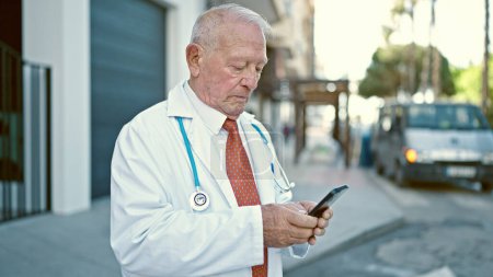 Téléchargez les photos : Homme âgé aux cheveux gris médecin debout avec une expression sérieuse à l'aide d'un smartphone dans la rue - en image libre de droit