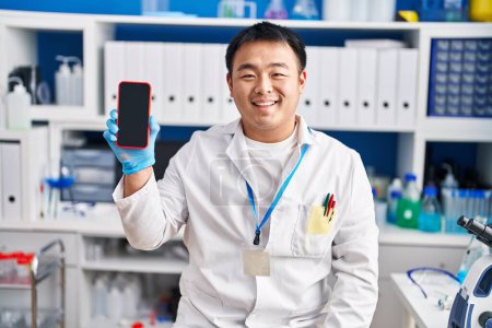 Téléchargez les photos : Jeune homme chinois travaillant au laboratoire scientifique tenant smartphone regardant positif et heureux debout et souriant avec un sourire confiant montrant les dents - en image libre de droit