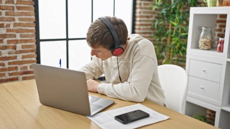 Téléchargez les photos : Jeune homme caucasien utilisant ordinateur portable et écouteurs écriture sur ordinateur portable à la salle à manger - en image libre de droit