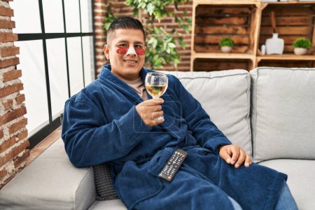 Téléchargez les photos : Jeune homme hispanique portant peignoir et sacs à paupières patchs boire du vin regardant positif et heureux debout et souriant avec un sourire confiant montrant des dents - en image libre de droit