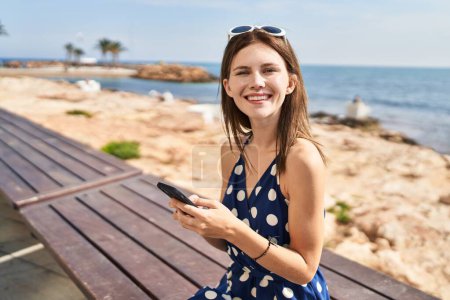 Téléchargez les photos : Jeune femme blonde touriste en utilisant smartphone assis sur le banc au bord de la mer - en image libre de droit