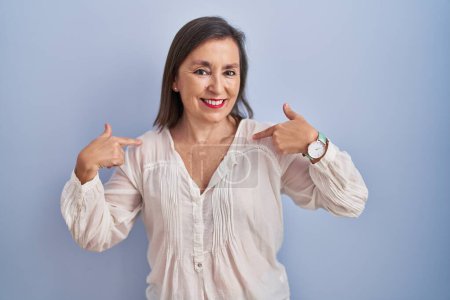 Foto de Mujer hispana de mediana edad de pie sobre fondo azul mirando confiado con sonrisa en la cara, señalándose con los dedos orgullosos y felices. - Imagen libre de derechos