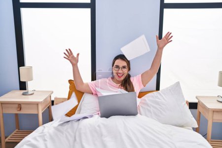 Photo for Young beautiful hispanic woman using laptop throwing paperwork sitting on bed at bedroom - Royalty Free Image