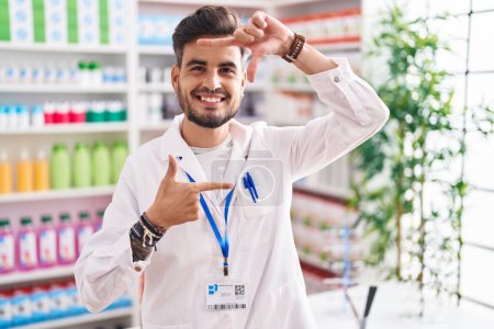 Téléchargez les photos : Jeune homme hispanique tatoué travaillant à la pharmacie pharmacie souriant faisant cadre avec les mains et les doigts avec le visage heureux. concept de créativité et photographie. - en image libre de droit