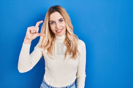 Téléchargez les photos : Jeune femme caucasienne debout sur fond bleu geste souriant et confiant avec la main faisant signe de petite taille avec les doigts regardant et la caméra. concept de mesure. - en image libre de droit