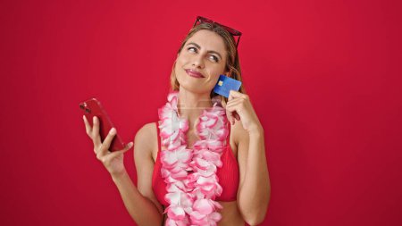 Foto de Mujer rubia joven que usa bikini de compras con teléfono inteligente y tarjeta de crédito pensando en el fondo rojo aislado - Imagen libre de derechos