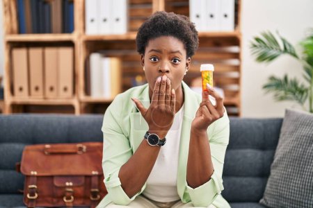 Foto de Mujer afroamericana que trabaja en la depresión sosteniendo píldoras que cubren la boca con la mano, conmocionada y temerosa de un error. expresión sorprendida - Imagen libre de derechos