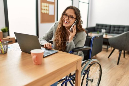 Photo for Young beautiful hispanic woman business worker talking on smartphone sitting on wheelchair at office - Royalty Free Image