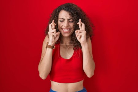 Téléchargez les photos : Femme hispanique avec les cheveux bouclés debout sur fond rouge geste doigt croisé souriant avec espoir et les yeux fermés. chance et concept superstitieux. - en image libre de droit