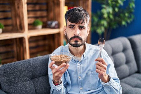 Foto de Joven hombre hispano con barba comiendo cereales integrales saludables relajado con expresión seria en la cara. simple y natural mirando a la cámara. - Imagen libre de derechos