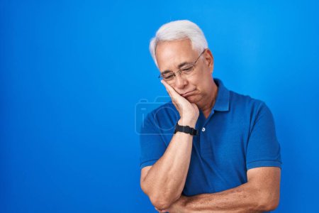 Téléchargez les photos : Homme d'âge moyen avec les cheveux gris debout sur fond bleu pensant à la recherche fatigué et ennuyé avec des problèmes de dépression avec les bras croisés. - en image libre de droit