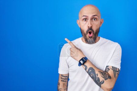 Foto de Hombre hispano con tatuajes de pie sobre fondo azul sorprendido señalando con el dedo hacia un lado, boca abierta expresión asombrada. - Imagen libre de derechos