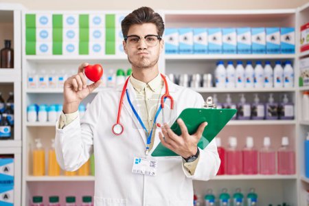 Téléchargez les photos : Young hispanic man working at pharmacy drugstore holding heart puffing cheeks with funny face. mouth inflated with air, catching air. - en image libre de droit