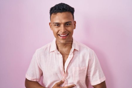Téléchargez les photos : Jeune homme hispanique debout sur fond rose souriant et riant fort à haute voix parce que drôle blague folle avec les mains sur le corps. - en image libre de droit