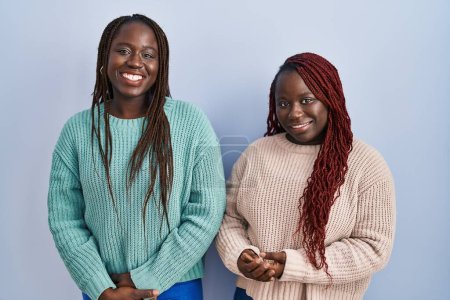 Téléchargez les photos : Deux femmes africaines debout sur fond bleu avec les mains jointes et les doigts croisés souriant détendu et joyeux. succès et optimisme - en image libre de droit
