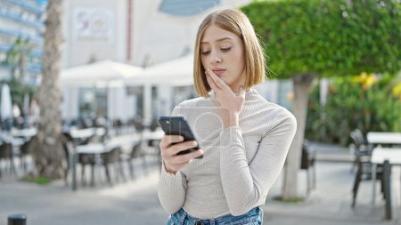 Téléchargez les photos : Jeune femme blonde utilisant un smartphone avec une expression sérieuse sur la terrasse du café - en image libre de droit