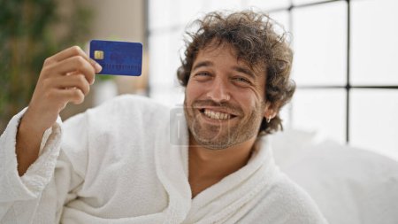 Photo for Young hispanic man wearing bathrobe holding credit card at bedroom - Royalty Free Image