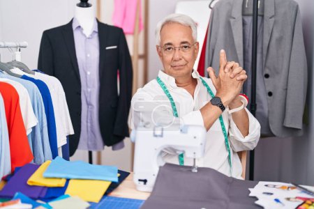 Photo for Middle age man with grey hair dressmaker using sewing machine holding symbolic gun with hand gesture, playing killing shooting weapons, angry face - Royalty Free Image
