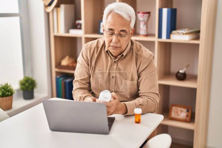 Photo for Senior man using laptop holding pills at home - Royalty Free Image