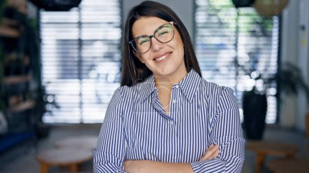Téléchargez les photos : Jeune belle femme hispanique portant des lunettes souriant heureux avec les bras croisés au bureau - en image libre de droit