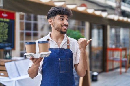 Photo for Arab man with beard wearing waiter apron at restaurant terrace pointing thumb up to the side smiling happy with open mouth - Royalty Free Image